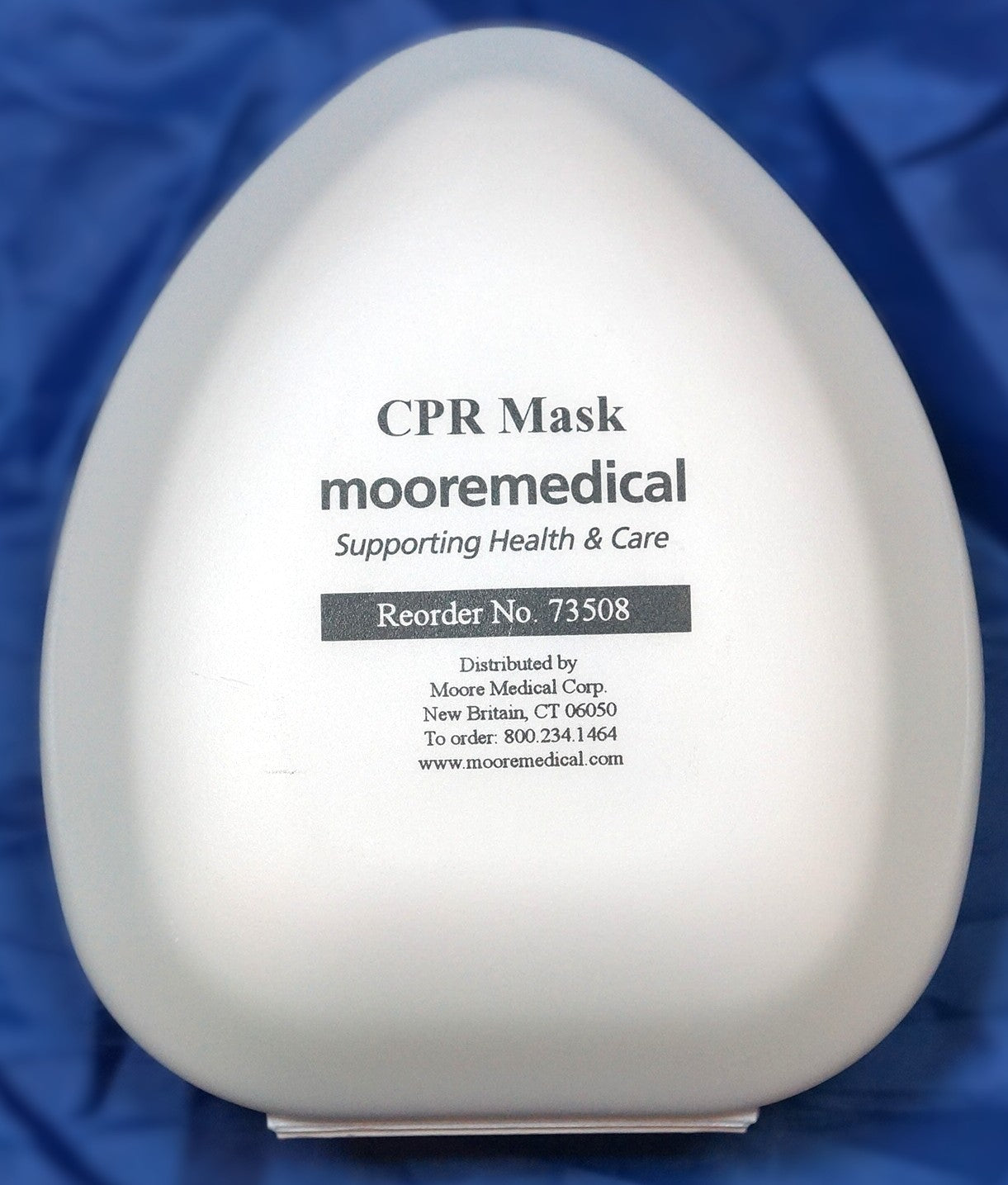 A white CPR mask container or packaging with printed text reading 'CPR Mask', 'mooremedical', and 'Supporting Health & Care', photographed against a blue fabric background.