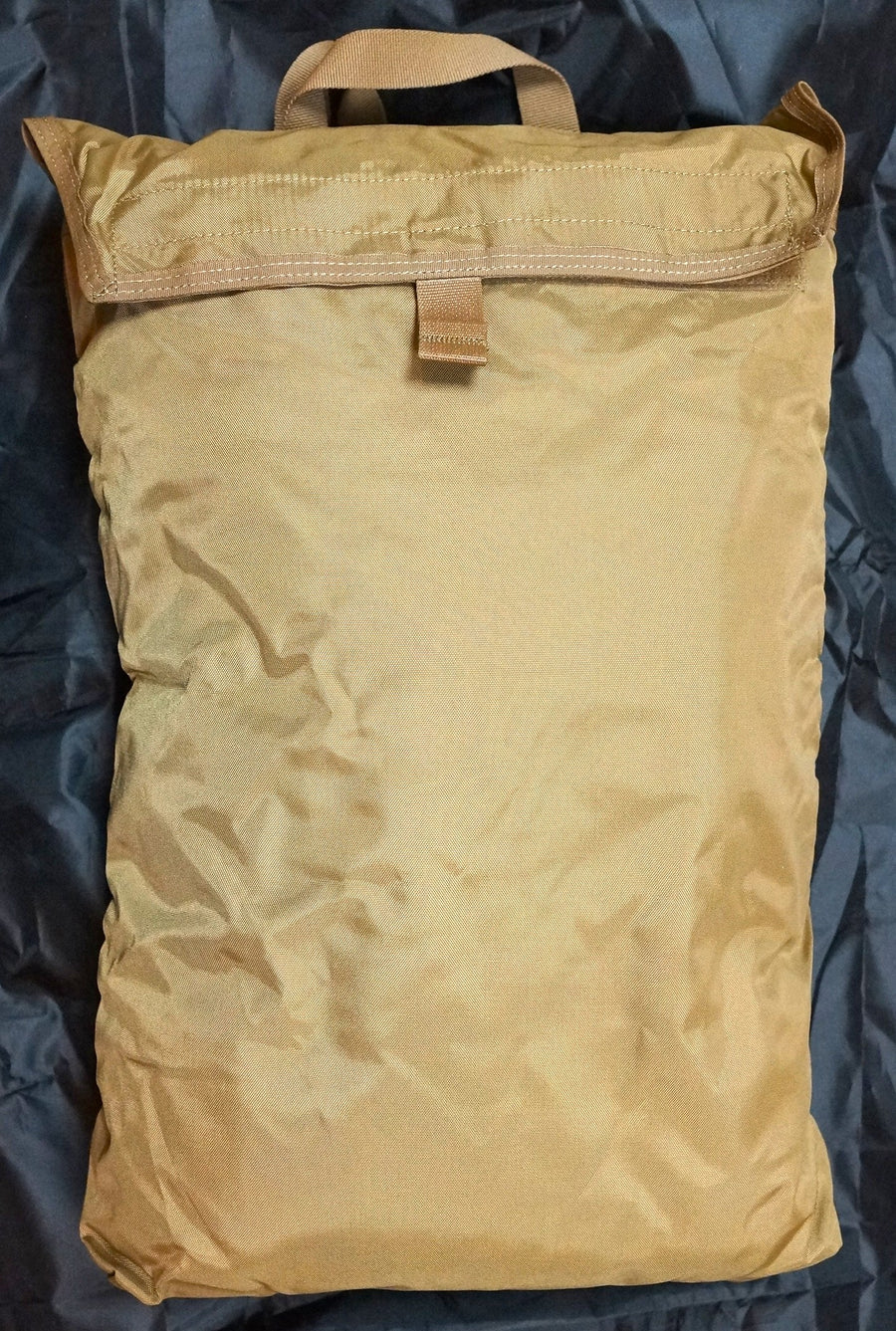 A tan nylon bag with a flap closure is shown next to a clear plastic bag containing a matching camouflage-patterned poncho liner, both laid flat on a black background.